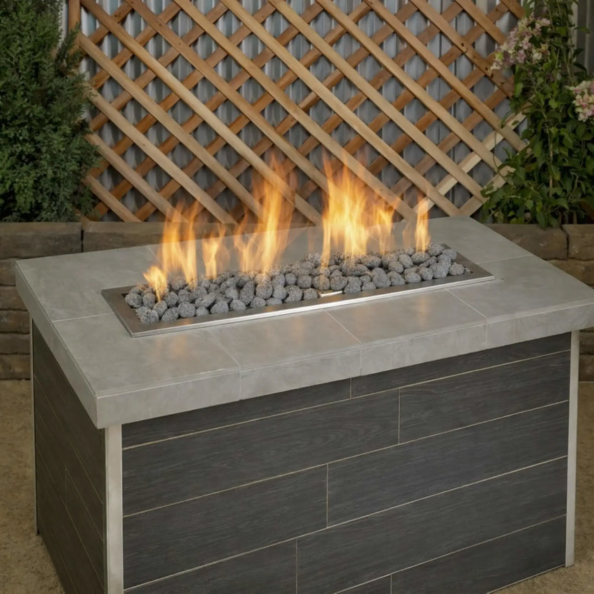 Rectangular outdoor gas fire table featuring small lava rocks evenly covering a stainless steel linear burner, with bright yellow flames against a wood lattice and garden backdrop.