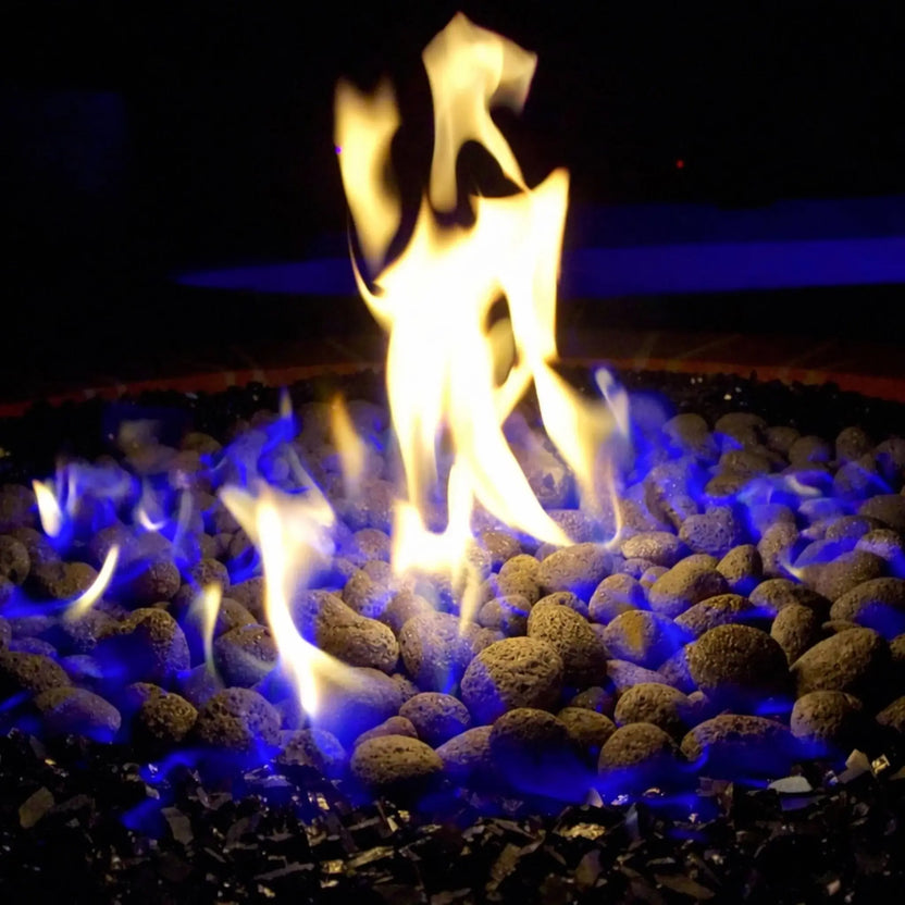 Close-up top view of a gas fire pit showing small lava rocks surrounded by black reflective fire glass, with vivid yellow flames and blue accents flickering above the burner.