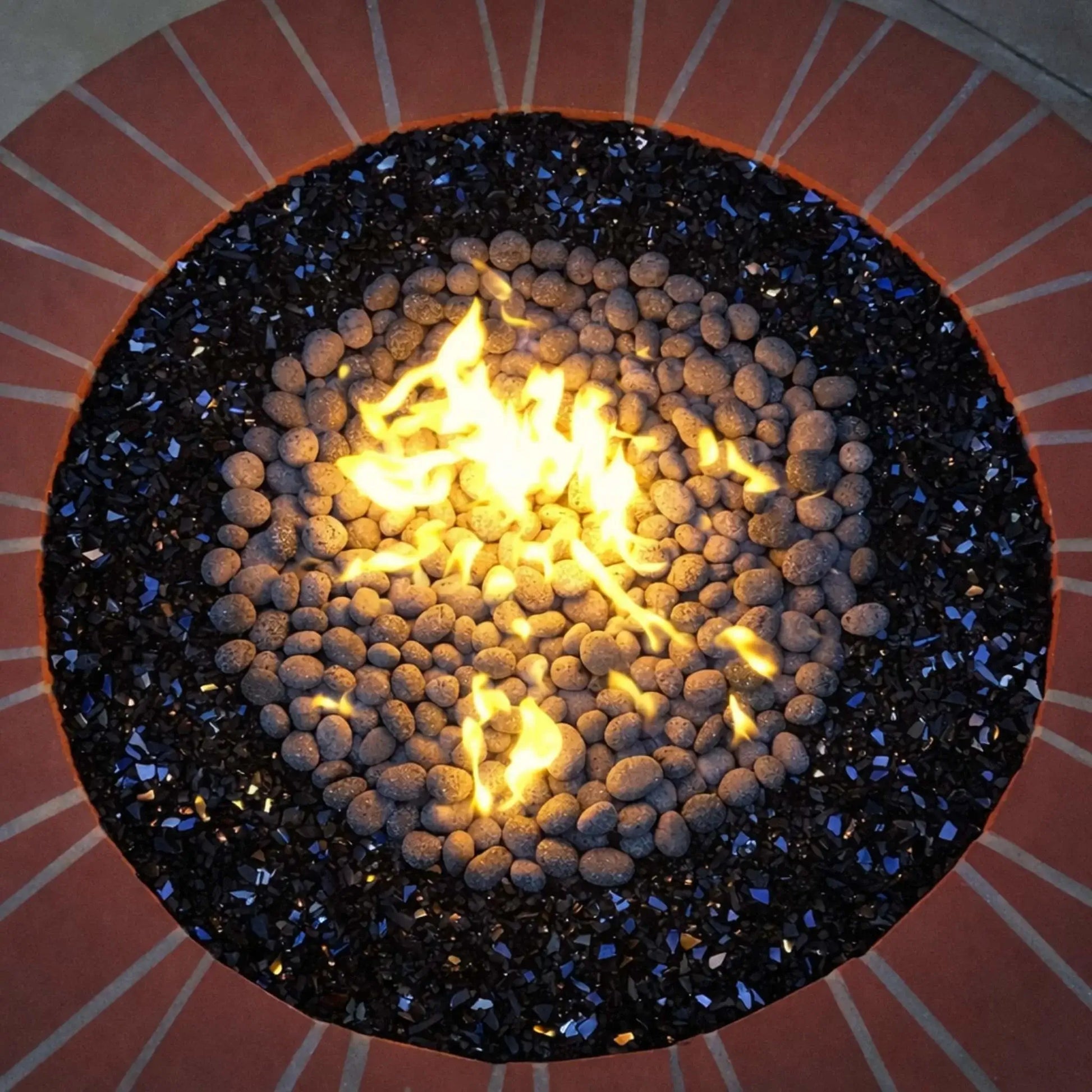 Overhead view of a round fire pit featuring small lava stones in the center, bordered by black reflective fire glass, with bright yellow flames rising from the middle.