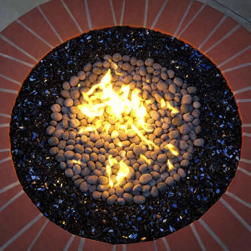Overhead view of a round fire pit featuring small lava stones in the center, bordered by black reflective fire glass, with bright yellow flames rising from the middle.