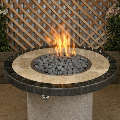 Round patio fire pit table topped with small gray lava stones encircling a central gas flame, set in a backyard setting with lattice fencing and surrounding greenery.