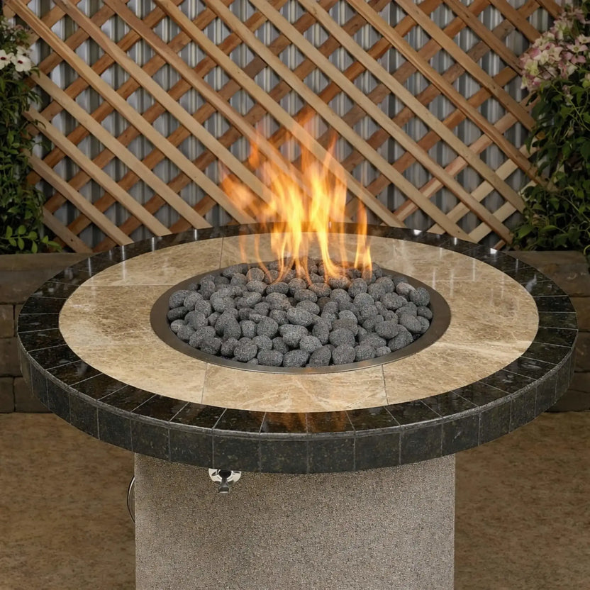 Round patio fire pit table topped with small gray lava stones encircling a central gas flame, set in a backyard setting with lattice fencing and surrounding greenery.