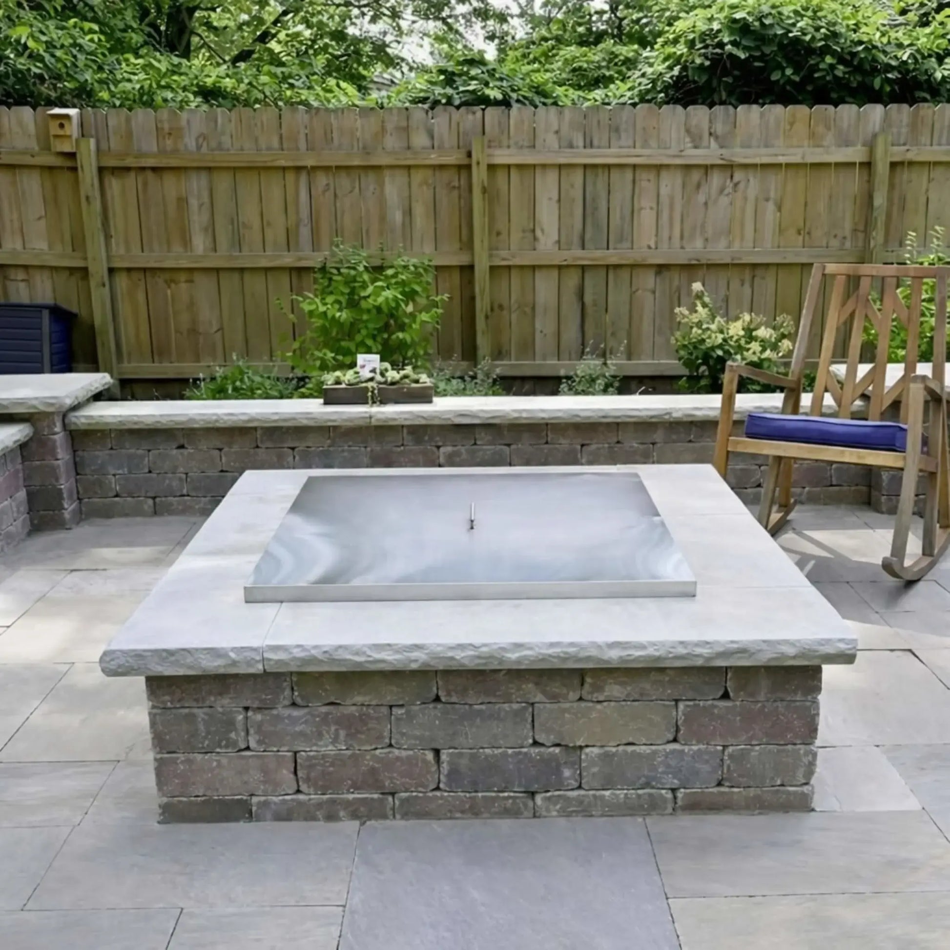 Square stainless steel fire pit burner cover installed flush inside a stone masonry fire pit, shown in a landscaped backyard patio with light gray pavers, retaining walls, and a wooden privacy fence.
