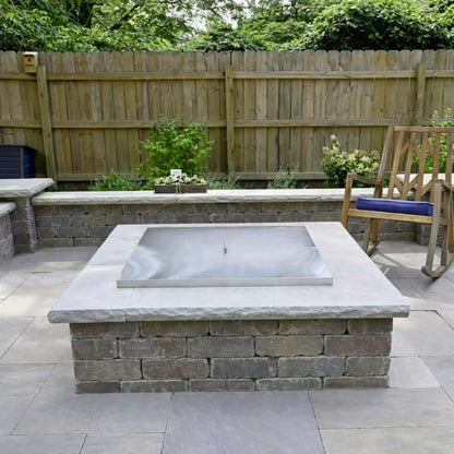 Square stainless steel fire pit burner cover installed flush inside a stone masonry fire pit, shown in a landscaped backyard patio with light gray pavers, retaining walls, and a wooden privacy fence.