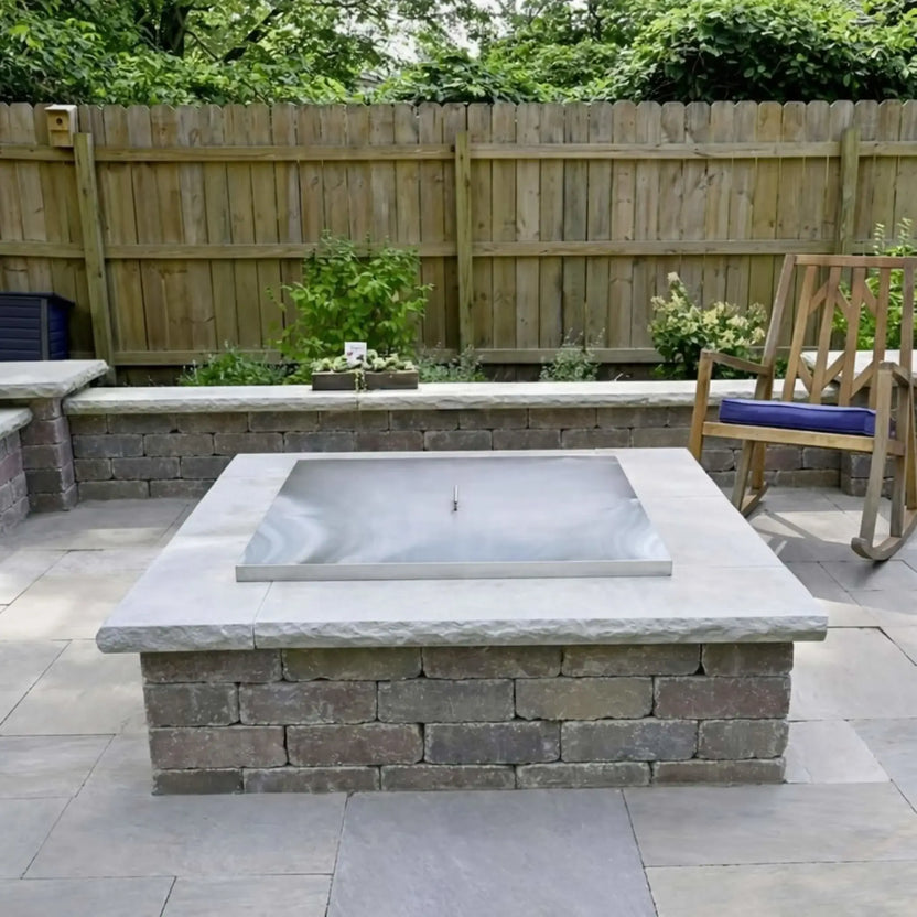 Square stainless steel fire pit burner cover installed flush inside a stone masonry fire pit, shown in a landscaped backyard patio with light gray pavers, retaining walls, and a wooden privacy fence.