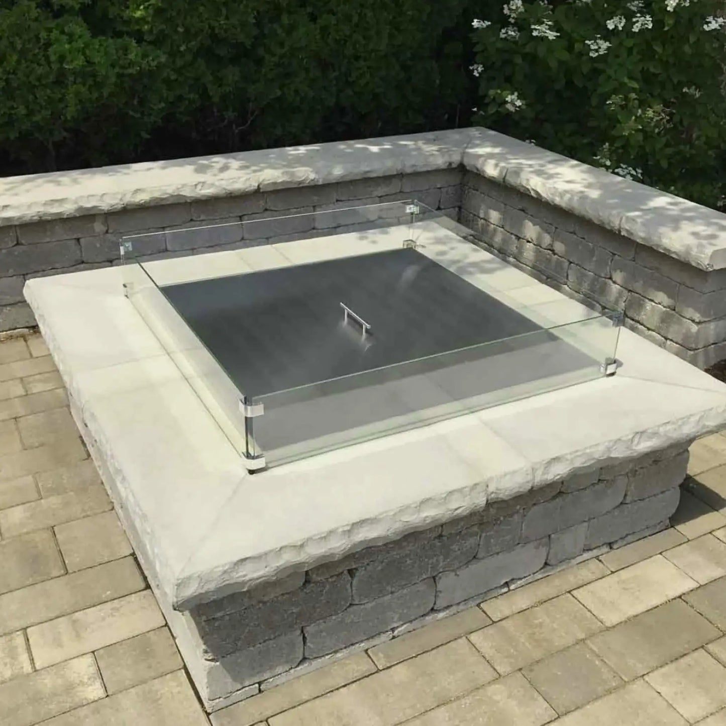 Square stainless steel fire pit burner cover resting on a stone fire pit with a clear glass wind guard installed around the perimeter, set on a paved patio surrounded by greenery and outdoor landscaping.