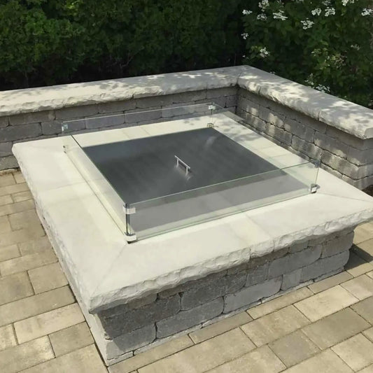 Square stainless steel fire pit burner cover resting on a stone fire pit with a clear glass wind guard installed around the perimeter, set on a paved patio surrounded by greenery and outdoor landscaping.