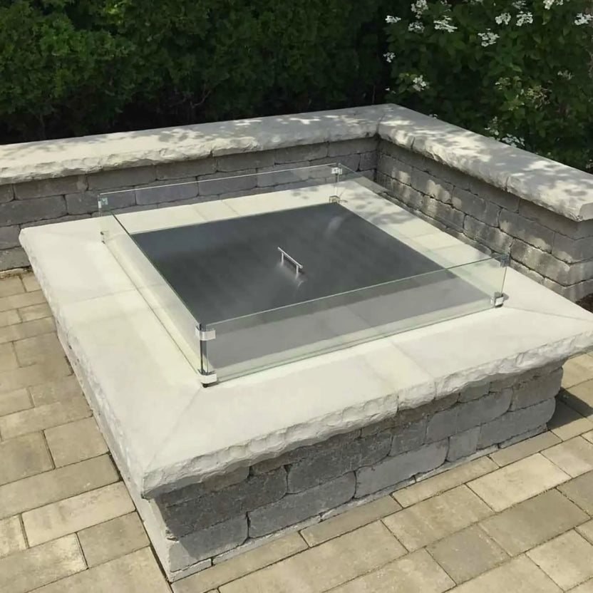 Square stainless steel fire pit burner cover resting on a stone fire pit with a clear glass wind guard installed around the perimeter, set on a paved patio surrounded by greenery and outdoor landscaping.