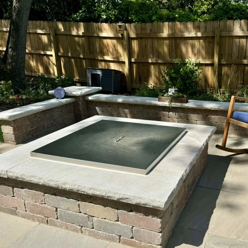 Outdoor square fire pit with a stainless steel burner cover placed over the burner opening, featured in a backyard seating area with stone walls, patio pavers, plants, and a wooden fence in natural daylight.