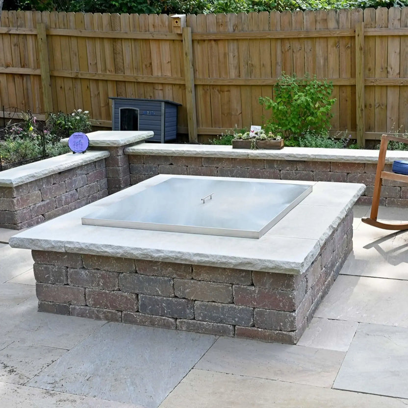 Stainless steel square fire pit burner cover used as a protective lid on a raised stone fire pit, shown in a finished outdoor living space with masonry walls, patio flooring, plants, and seating nearby.