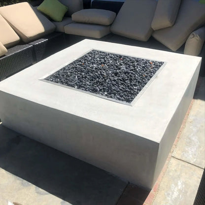 Square concrete fire pit with a stainless steel drop-in burner pan filled with black lava rock, set into a modern outdoor seating area with woven sectional furniture and neutral cushions.