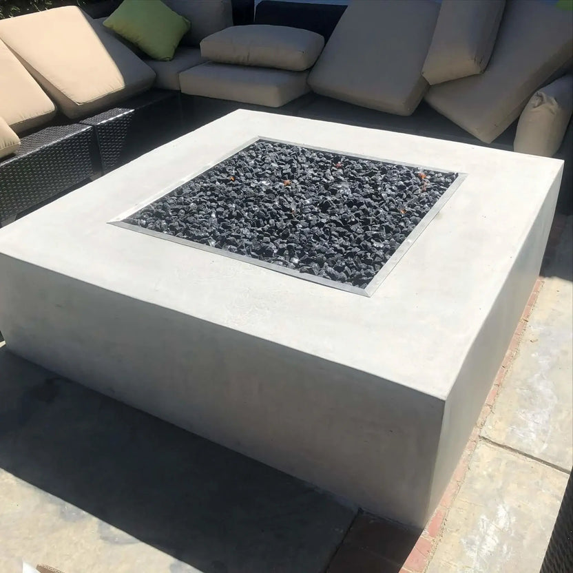 Square concrete fire pit with a stainless steel drop-in burner pan filled with black lava rock, set into a modern outdoor seating area with woven sectional furniture and neutral cushions.