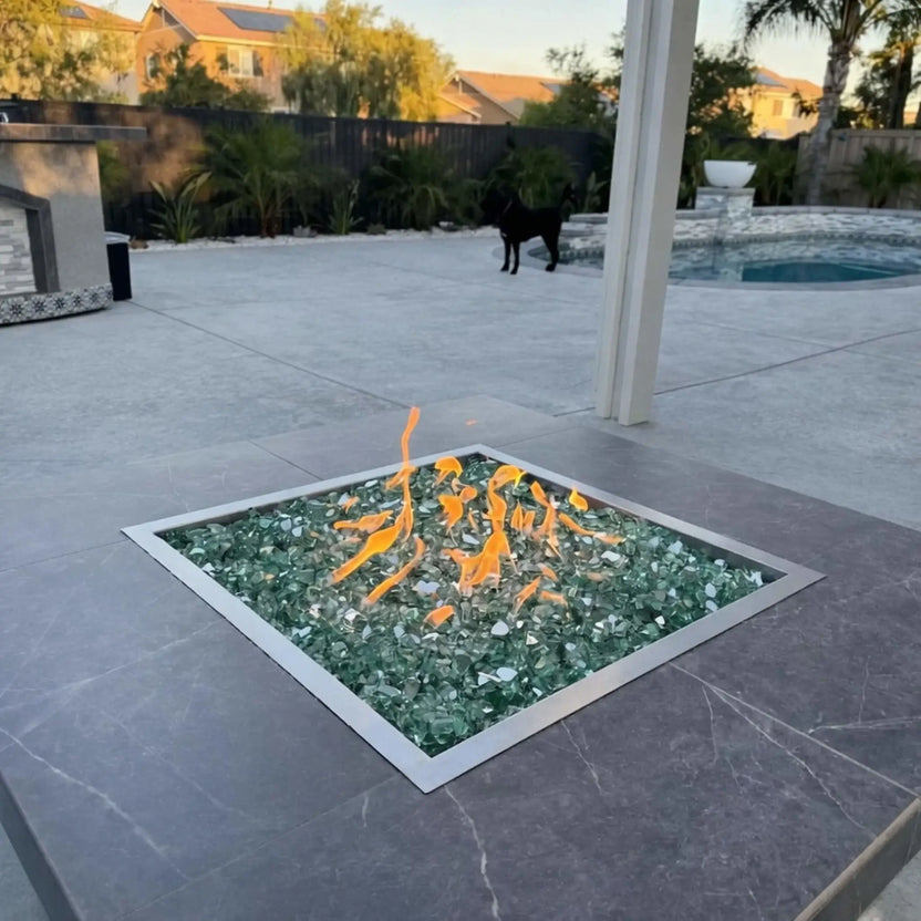 Close-up view of a square stainless steel fire pit burner pan with green fire glass and active flames, installed in a stone-topped outdoor fire pit near a pool and patio area.