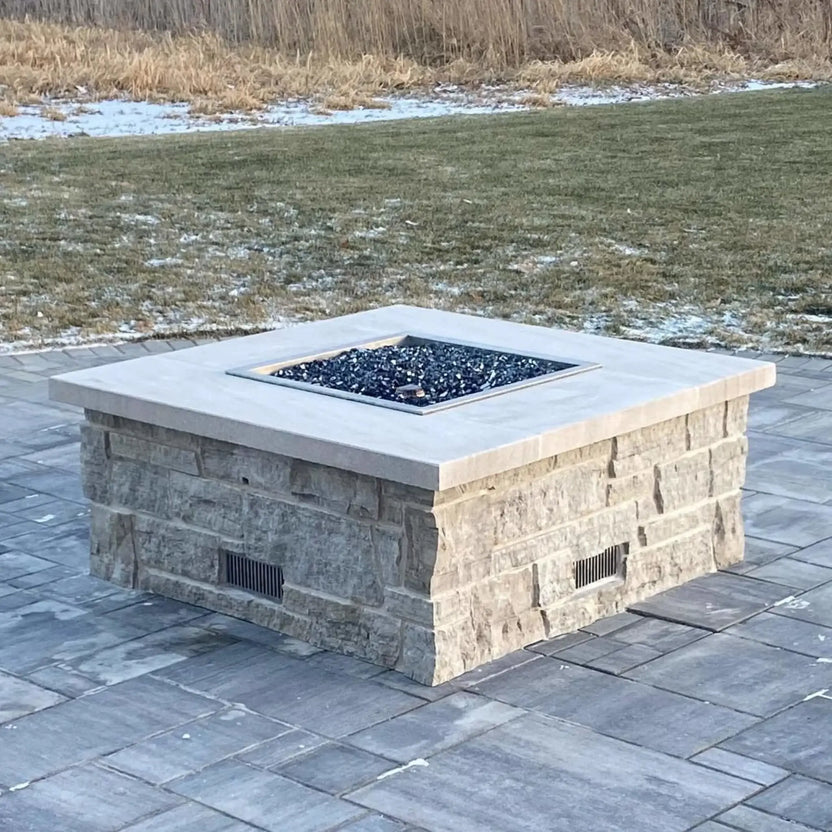 Square fire pit burner pan with shimmering green fire glass and tall flames, set into a modern stone fire pit overlooking a landscaped backyard and pool area.