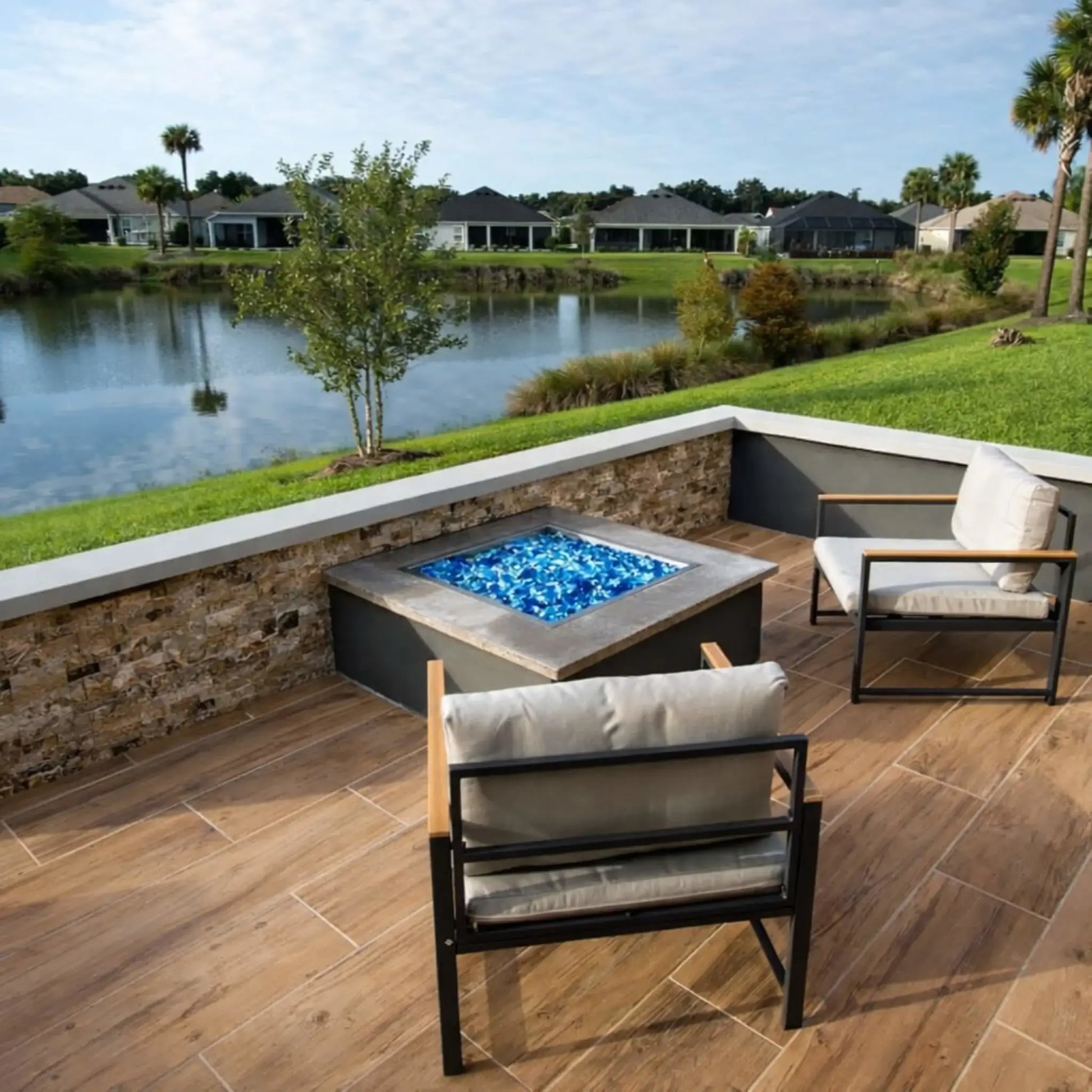 Custom stone fire pit featuring a square stainless steel drop-in burner pan filled with black fire glass, positioned on a paved patio with winter grass and natural surroundings.