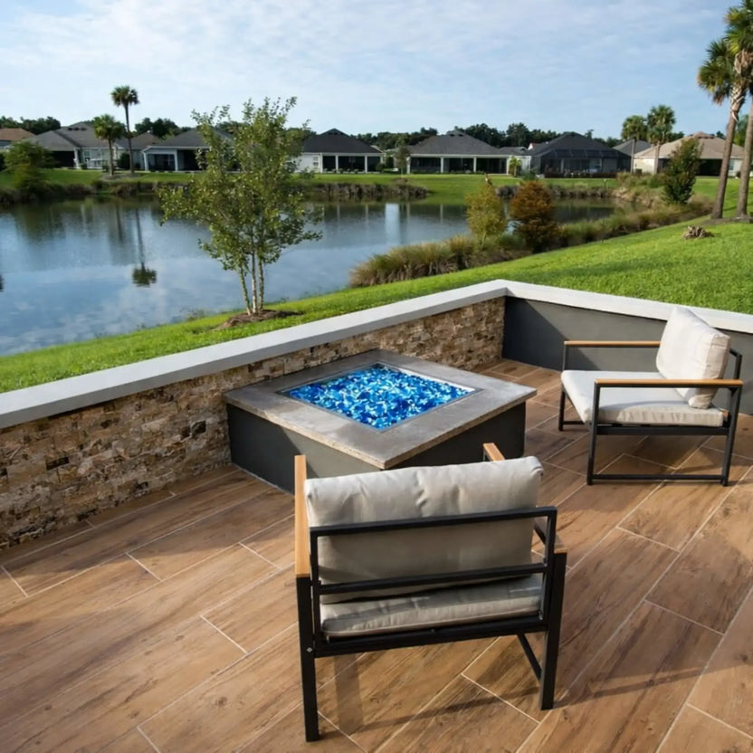 Custom stone fire pit featuring a square stainless steel drop-in burner pan filled with black fire glass, positioned on a paved patio with winter grass and natural surroundings.