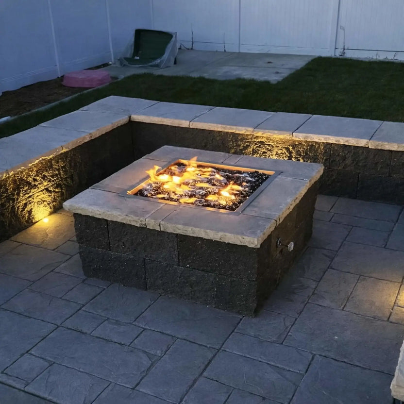 Outdoor fire pit built with stacked stone and a square stainless steel burner pan, accented with blue fire glass and placed on a raised patio overlooking a lakefront community.