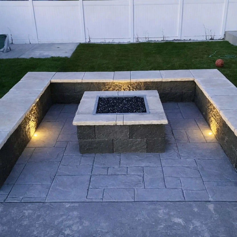 Evening backyard fire pit scene showing a square stainless steel burner pan with flames and fire glass, integrated into a seating wall with built-in accent lighting.