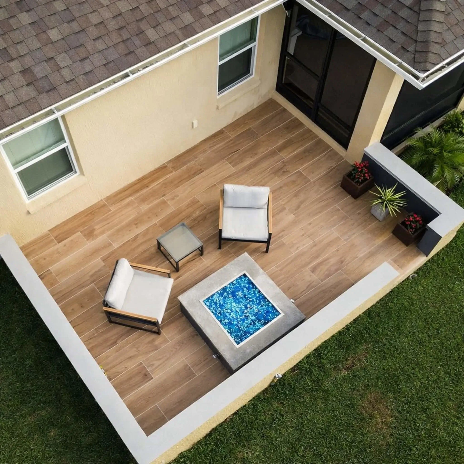 Overhead view of a modern patio featuring a square fire pit table with blue fire glass, stainless steel burner pan, and minimalist outdoor chairs arranged on wood-look tile flooring.