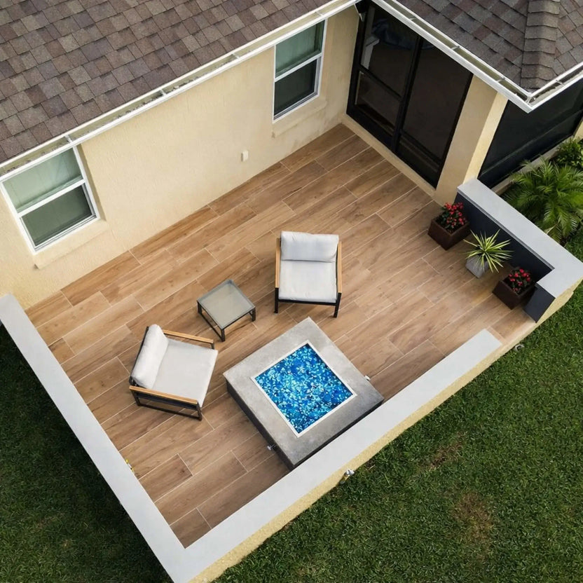 Overhead view of a modern patio featuring a square fire pit table with blue fire glass, stainless steel burner pan, and minimalist outdoor chairs arranged on wood-look tile flooring.