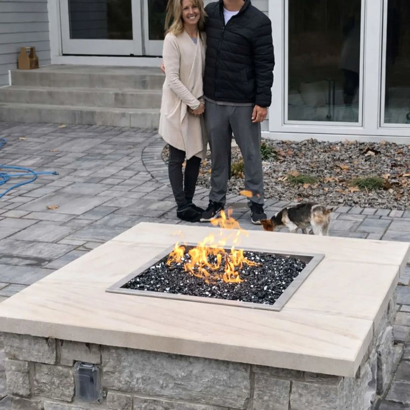 Homeowners standing beside a custom stone fire pit with a square stainless steel burner pan and black fire glass, flames burning steadily in a finished backyard patio setting.