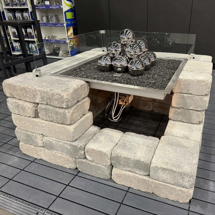 Square outdoor fire pit burning at night with stainless steel burner pan and gas logs, set on a paved patio with light snow on the ground and a warmly lit home in the background.