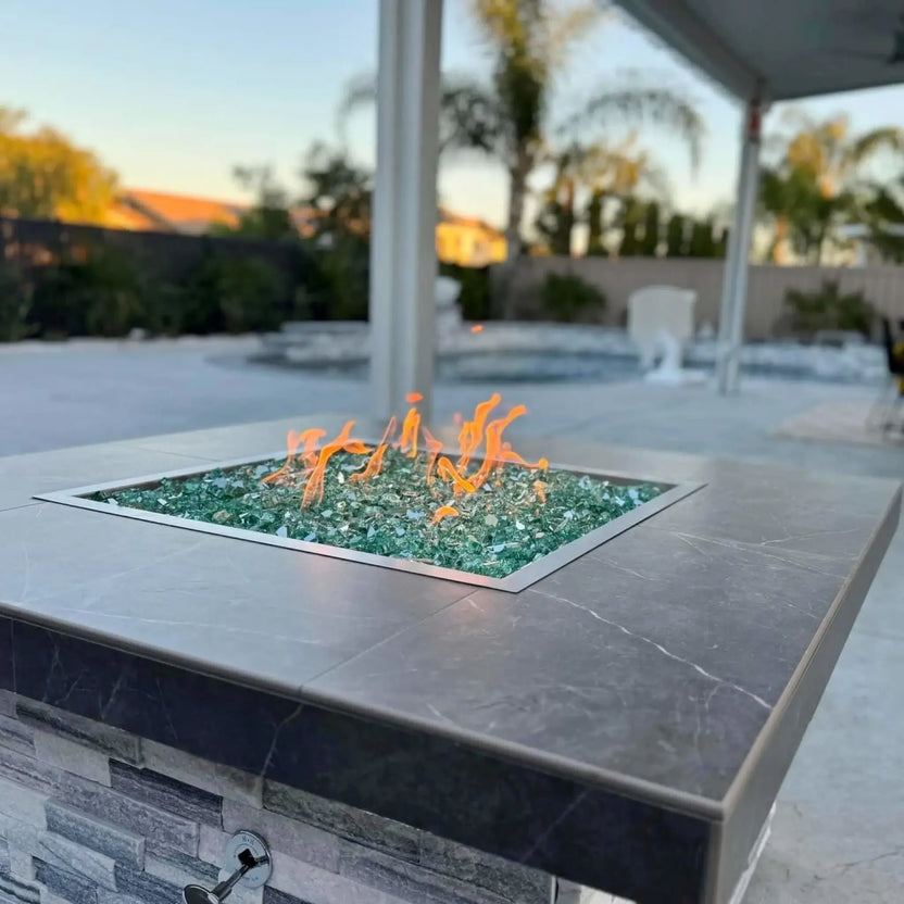 Square stone fire pit featuring a stainless steel drop-in burner pan filled with decorative fire glass and polished fire spheres, displayed in an indoor showroom setting.