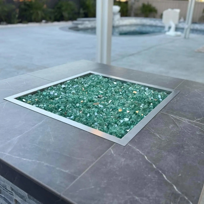 Modern outdoor square fire pit with stainless steel burner pan and green fire glass, producing vibrant flames on a covered patio with a pool area in the background.