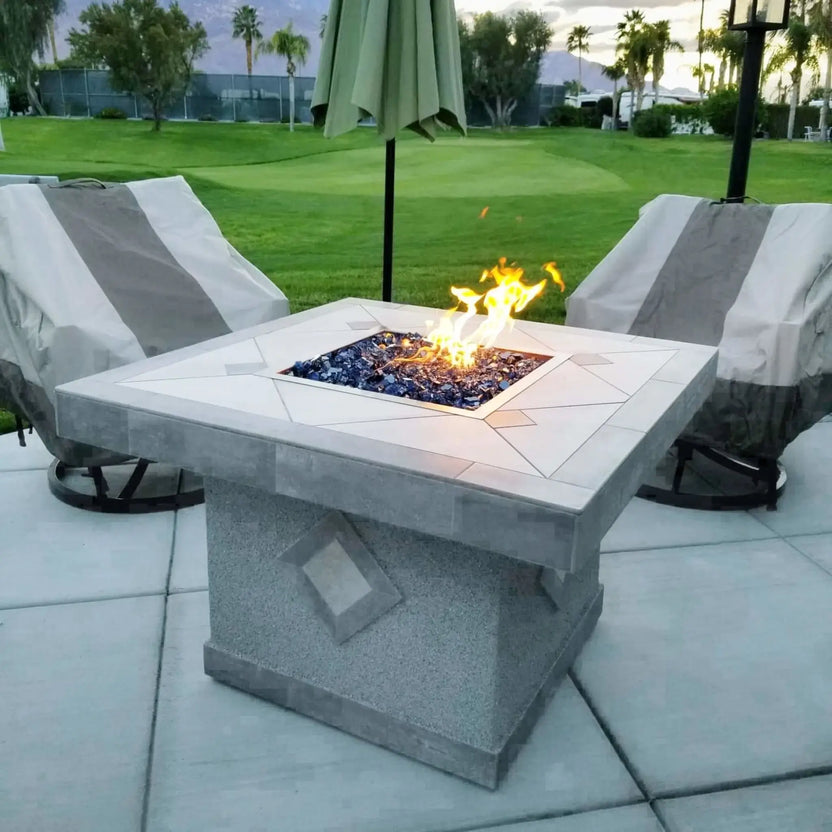 Square fire pit table with stainless steel drop-in burner pan and blue fire glass, positioned on a patio overlooking a manicured green lawn and golf course scenery.