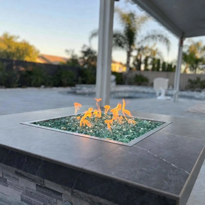 Close-up of a square stainless steel fire pit burner pan filled with green fire glass, showing clean flame pattern and flush installation within a stone fire pit table.