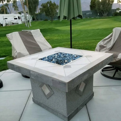 Square outdoor fire pit with stainless steel burner pan filled with blue fire glass, centered between cushioned patio chairs beneath an outdoor umbrella in a landscaped backyard.