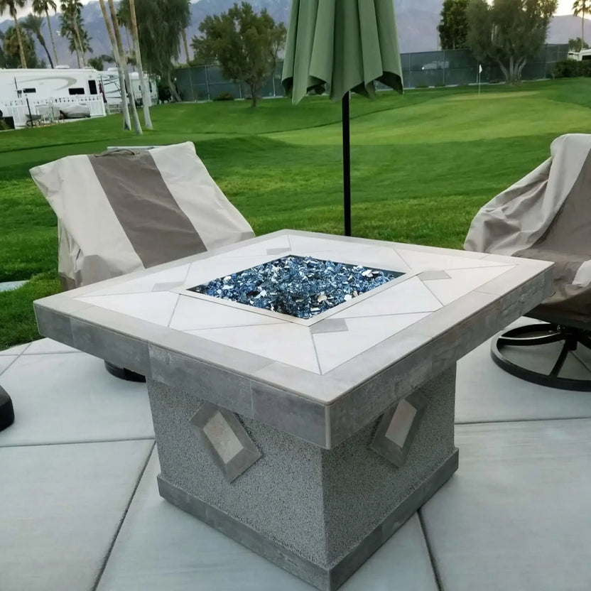 Square outdoor fire pit with stainless steel burner pan filled with blue fire glass, centered between cushioned patio chairs beneath an outdoor umbrella in a landscaped backyard.