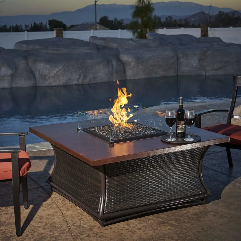 Square tempered glass wind guard installed on a modern outdoor fire table, surrounding a lit fire feature and blending seamlessly with patio furniture in a backyard setting.