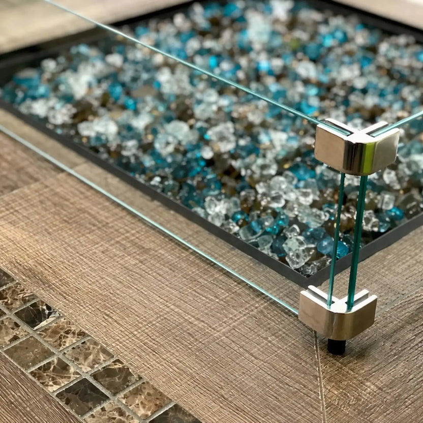 Detailed view of a square tempered glass wind guard corner bracket securing clear glass panels around a fire pit filled with mixed blue fire glass on a textured tabletop surface.