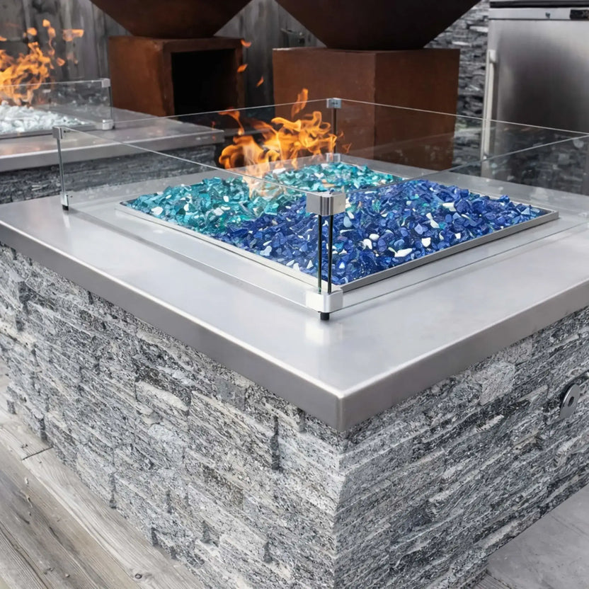 Square outdoor fire pit with tempered glass wind guard and blue fire glass, built into a stone surround with a clean, modern look in an open-air backyard setting.