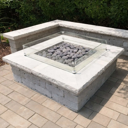 Square fire pit enclosure with a clear glass wind guard and smooth gray lava stones, set into a stone masonry base on a paved patio surrounded by greenery.