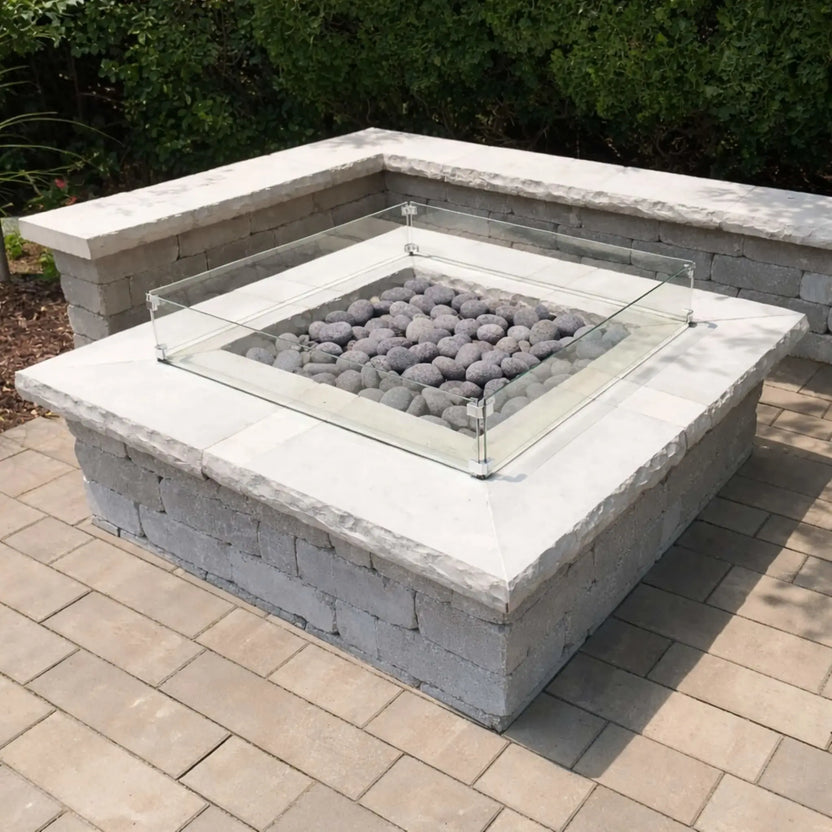 Square fire pit enclosure with a clear glass wind guard and smooth gray lava stones, set into a stone masonry base on a paved patio surrounded by greenery.