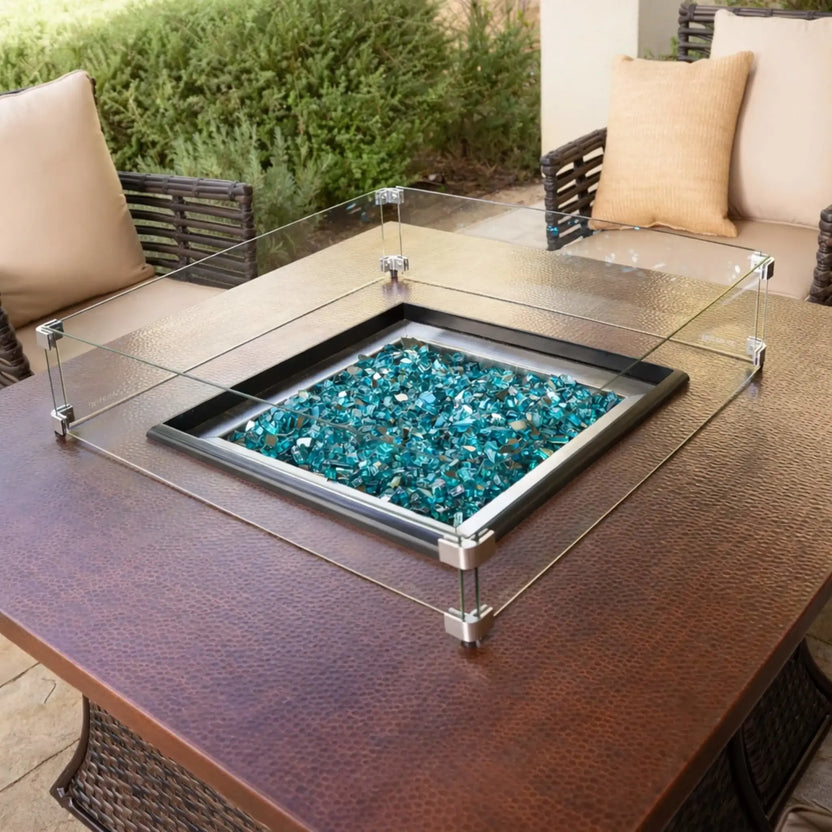Square fire pit table featuring a tempered glass wind guard and blue fire glass, styled with wicker patio chairs and neutral cushions in a landscaped outdoor seating area.