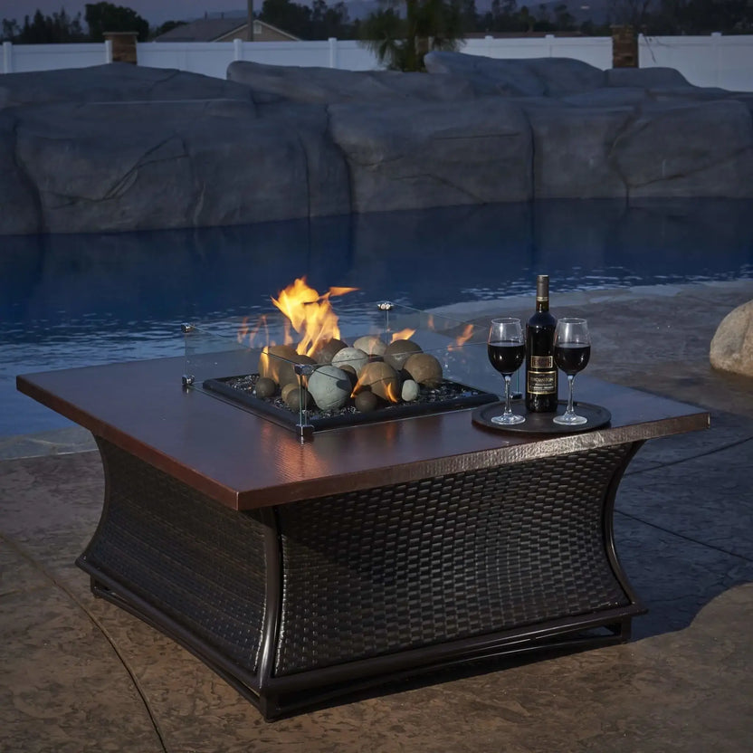 Outdoor fire pit table featuring a square glass wind guard enclosing decorative fire media, shown in a stylish patio environment with seating and evening ambiance.