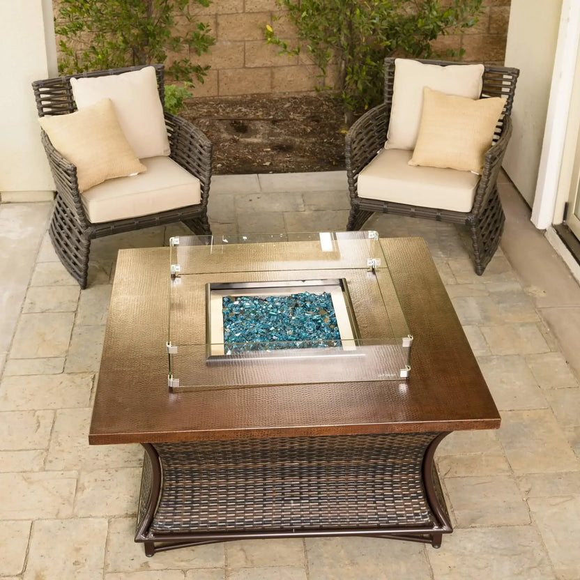Square fire pit with clear tempered glass wind guard protecting active flames, displayed on a woven outdoor fire table with decorative fire glass in a cozy backyard space.