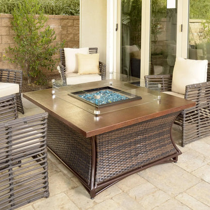 Square outdoor fire pit table with a tempered glass wind guard surrounding blue fire glass, set on a covered patio with woven wicker seating, neutral cushions, and a warm bronze tabletop finish.