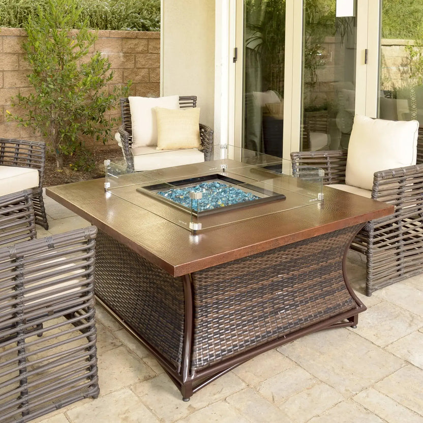 Square outdoor fire pit table with a tempered glass wind guard surrounding blue fire glass, set on a covered patio with woven wicker seating, neutral cushions, and a warm bronze tabletop finish.