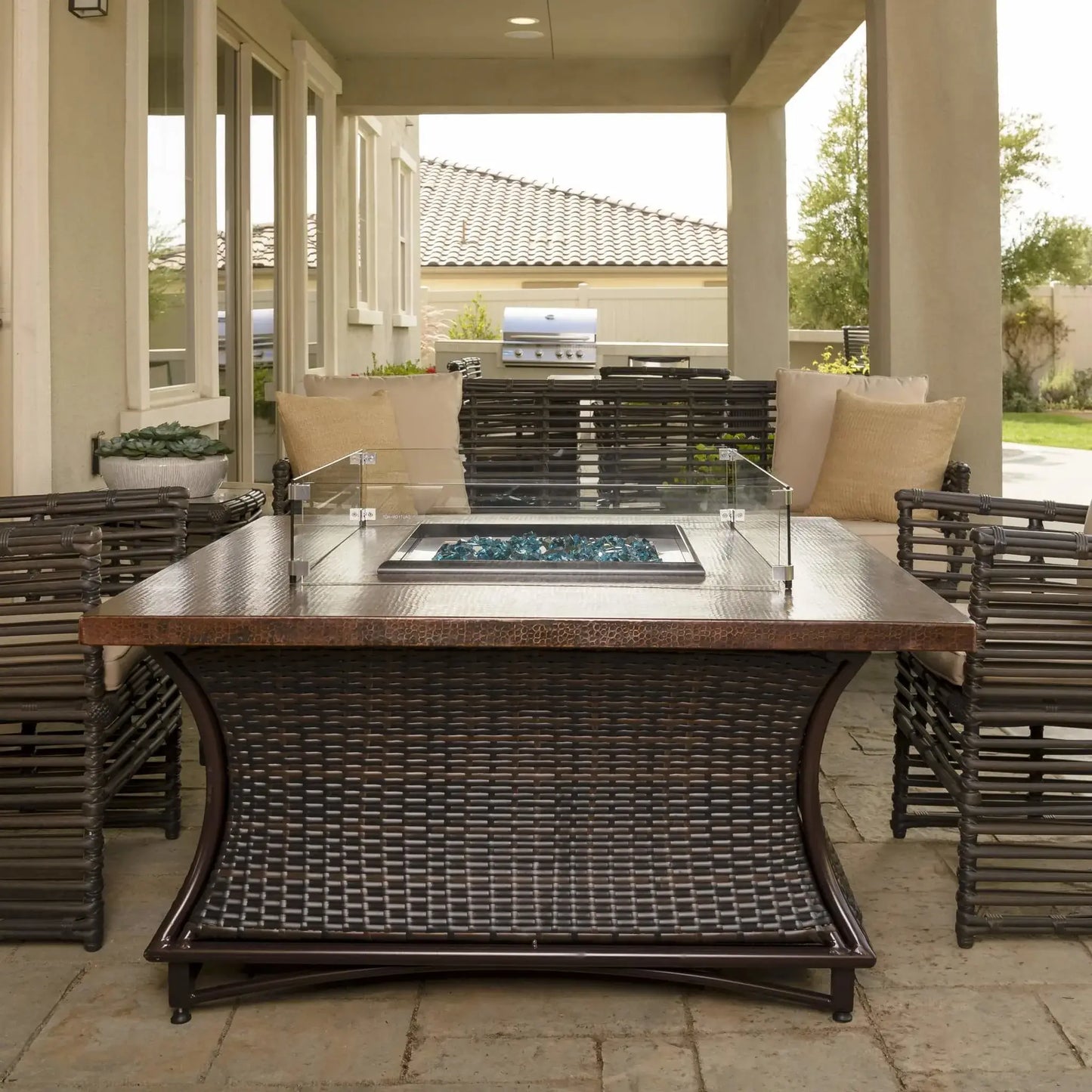Front view of a square fire pit table featuring a clear glass wind guard and blue fire glass, positioned under a covered outdoor living space with cushioned wicker chairs and a built-in grill in the background.