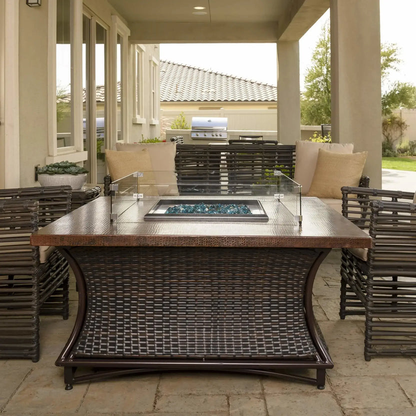 Front view of a square fire pit table featuring a clear glass wind guard and blue fire glass, positioned under a covered outdoor living space with cushioned wicker chairs and a built-in grill in the background.