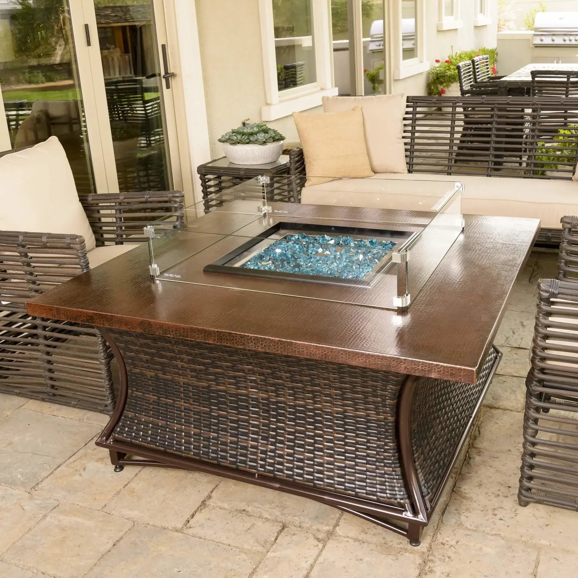 Angled view of a square gas fire pit table with tempered glass wind guard and reflective blue fire glass, surrounded by woven patio seating and decorative planters on a stone paver patio.