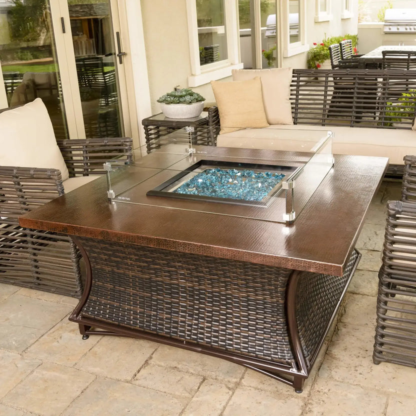 Angled view of a square gas fire pit table with tempered glass wind guard and reflective blue fire glass, surrounded by woven patio seating and decorative planters on a stone paver patio.