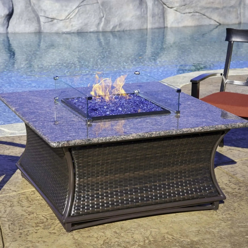 Square fire pit table with a raised glass wind guard and blue fire glass burning over a granite-style surface, placed beside a swimming pool with outdoor lounge seating nearby.