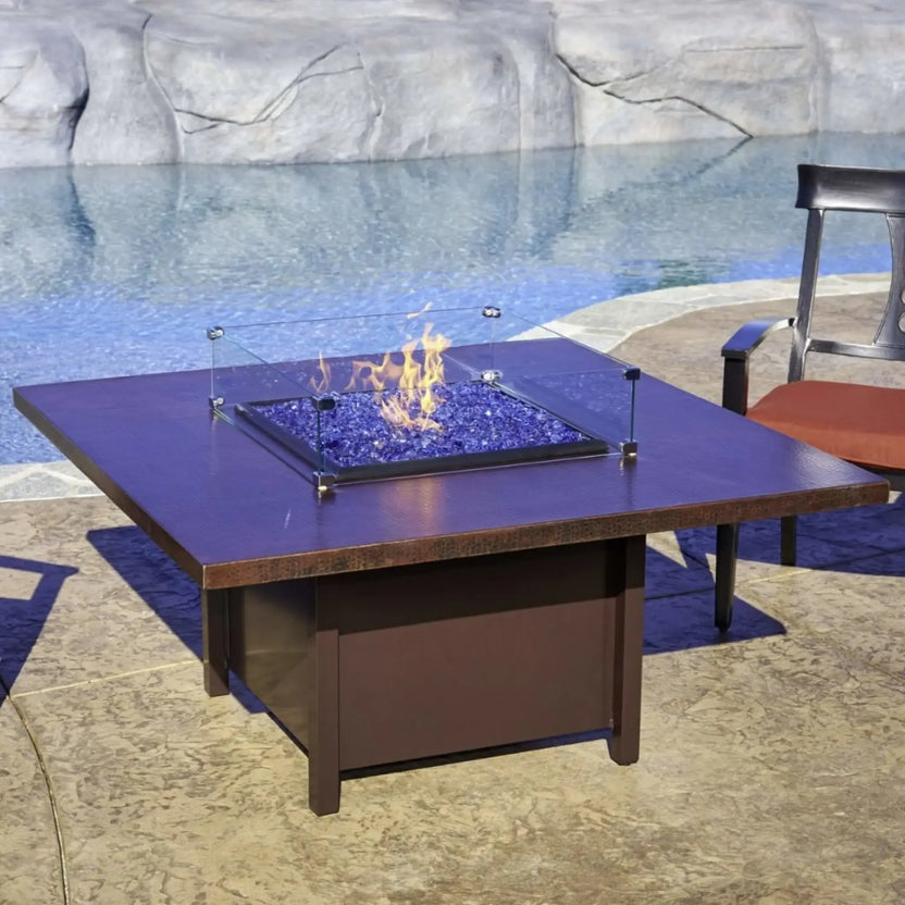 Modern square fire pit table with tempered glass wind guard enclosing blue fire glass flames, positioned poolside on a textured stone patio with outdoor chairs in the background.