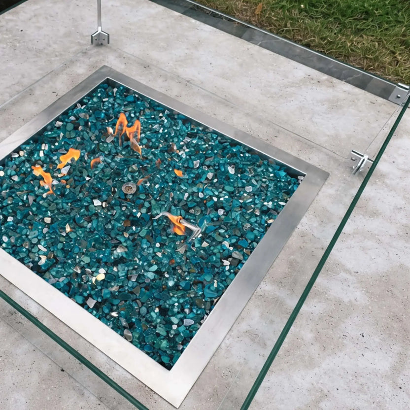 Close-up of a square fire pit burner filled with blue fire glass and active flames, protected by a clear tempered glass wind guard mounted with metal corner brackets.