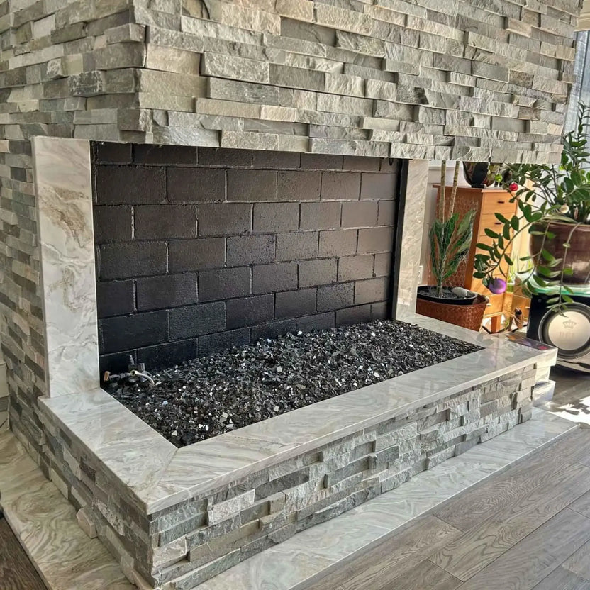 Modern indoor gas fireplace featuring a stainless steel H-style burner filled with black and gray fire glass, set inside a stacked stone surround with a dark firebrick interior, showcasing a clean, contemporary fireplace design ready for ignition.